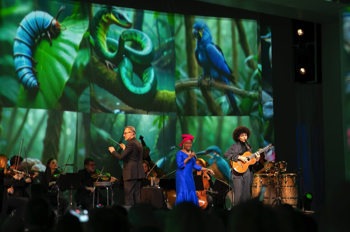 Angélique Kidjo and Will Santt playing together with the orchestra behind them against a back drop of projected nature images generated by AI created by Refik Anadol.