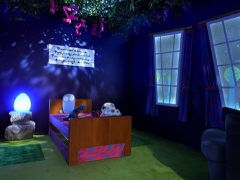 The 100 Story Hotel Dinosaur Bedroom. The little dinosaur has insomnia and peeks out from under the blanket of his bed. Moonlight streams through the vines across the ceiling and across the pink hanging Z's over the dinosaur's bed. A glowing dinosaur egg sits on the bedside table along with a phone to reception.