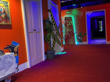 A photo of the central hallway in the 100 Story hotel. A penguin porter pushes a room service trolley. A hazard-taped door has fluorescent green goo seeping out from below the door. At the end of the hall is a glowing door leading to the disco room number 1999.