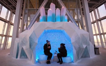 Two people sit inside the iridescent blue crystal grotto wrapped up in coats drinking hot drinks.