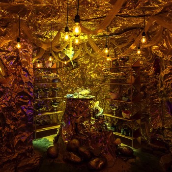  The treasure room, completely clad in gold with a central plinth with a tray of antiquities. Exposed filament light bulbs hang from the ceiling. 